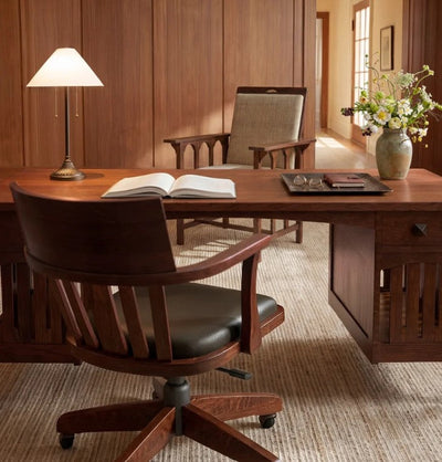 A warm wood office scene with a large wooden desk, a dark wood swivel chair with black leather seating, an open book on the desk, a table lamp, and a vase of flowers against wood-paneled walls.