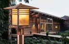A close-up of a warm glowing outdoor lantern post in the foreground, with a modern wooden house and garden visible in the softly blurred background.
