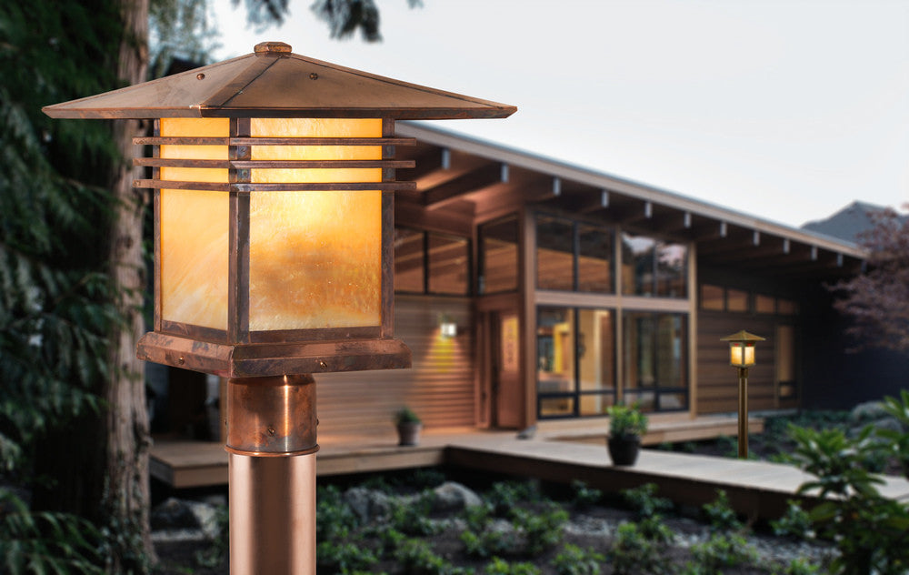 A close-up of a warm glowing outdoor lantern post in the foreground, with a modern wooden house and garden visible in the softly blurred background.
