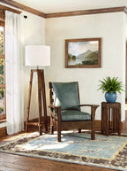 A styled interior corner featuring a dark green leather armchair with a wooden frame, a tall wooden tripod floor lamp, a small wooden side table with a potted fern, and a patterned area rug on a hardwood floor.