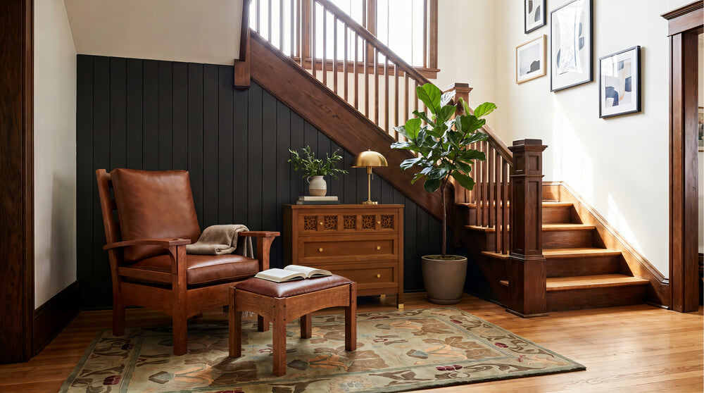 A styled interior corner featuring a brown leather armchair with a matching wooden footstool, set on a patterned area rug beside a wooden dresser and next to a staircase with dark paneled walls.