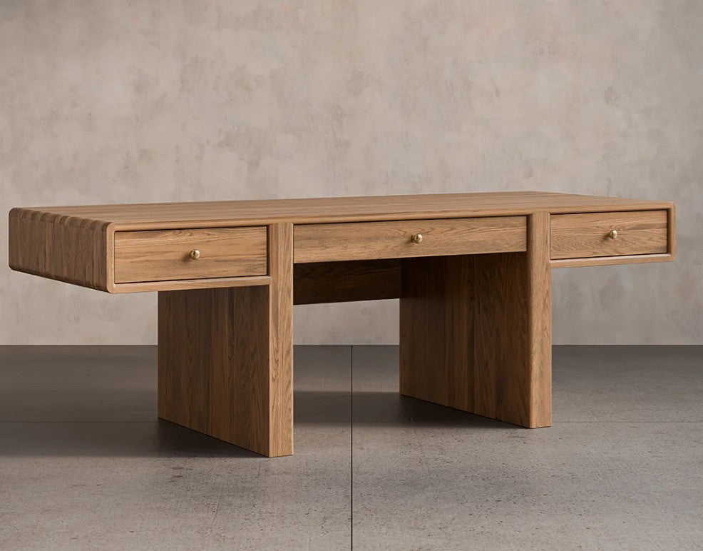 A mid-century modern oak wood writing desk with three drawers, featuring brass drawer pulls, set against a textured concrete wall and floor.