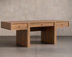 A mid-century modern oak wood writing desk with three drawers, featuring brass drawer pulls, set against a textured concrete wall and floor.