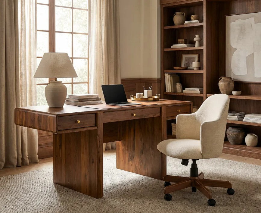 A warm-toned home office scene featuring a dark wood writing desk with drawers, a cream upholstered swivel chair, a table lamp, books, and a laptop. A tall built-in wooden bookshelf stands behind the desk in a styled room with beige curtains and neutral decor.