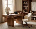 A warm-toned home office scene featuring a dark wood writing desk with drawers, a cream upholstered swivel chair, a table lamp, books, and a laptop. A tall built-in wooden bookshelf stands behind the desk in a styled room with beige curtains and neutral decor.
