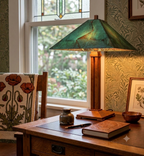 A vintage-style table lamp with a textured green glass conical shade and a warm wood base sits on a wooden desk in a cozy room setting with wallpaper, a window, and decorative items.