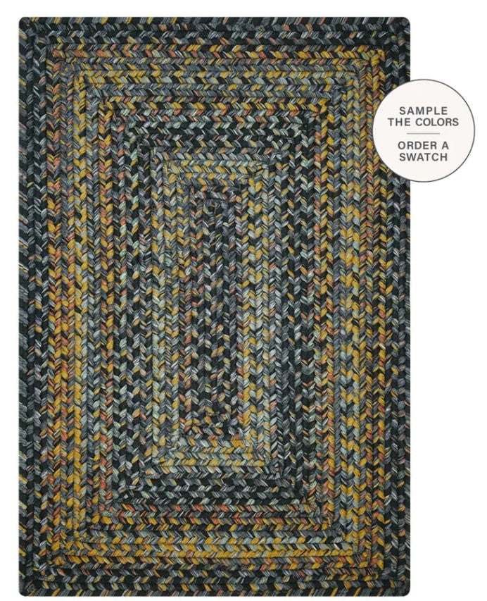 A braided rectangular area rug with concentric borders in a textured herringbone pattern, featuring dark gray, black, mustard yellow, and beige tones. A white circular badge in the top right corner reads 'SAMPLE THE COLORS ORDER A SWATCH'.