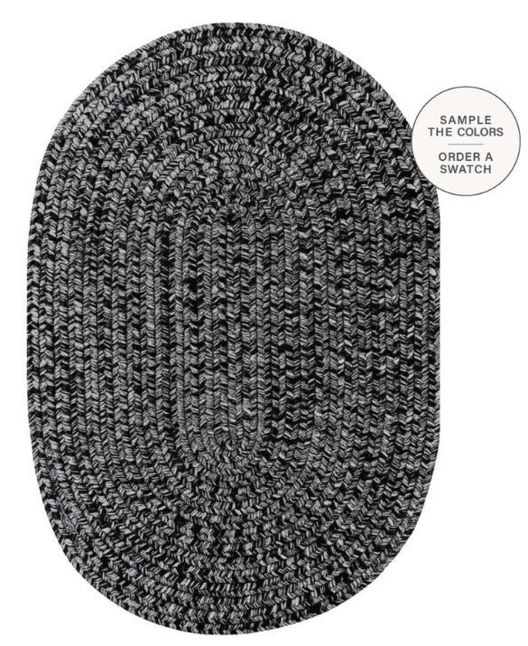 An oval braided area rug in a black and white heathered pattern, shown from a top-down view on a white background with a circular text overlay in the top right that reads 'SAMPLE THE COLORS ORDER A SWATCH'.