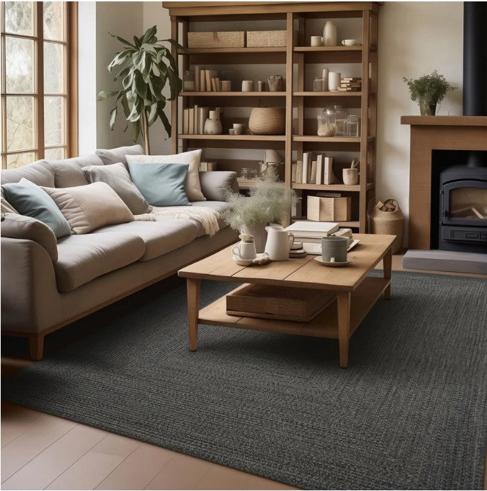 A large charcoal gray textured area rug in a cozy, modern living room setting with a beige sofa, wooden coffee table, and a tall wooden bookshelf.