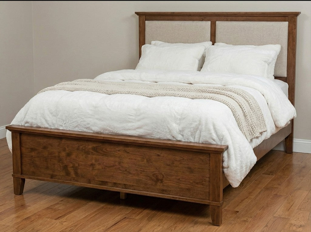 A medium brown wooden platform bed frame with a tall upholstered headboard and footboard, shown dressed with white bedding and a cream knit throw on a hardwood floor against a beige wall.