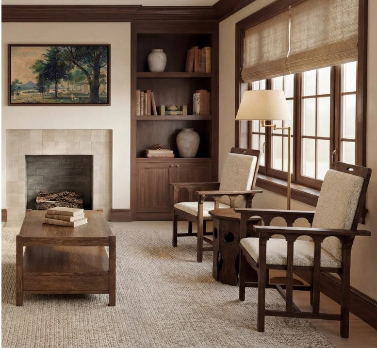 A cozy living room scene featuring two dark wood armchairs with cream upholstered seats, a matching wooden coffee table, a floor lamp, a built-in wooden bookshelf, and a fireplace with a landscape painting above it.