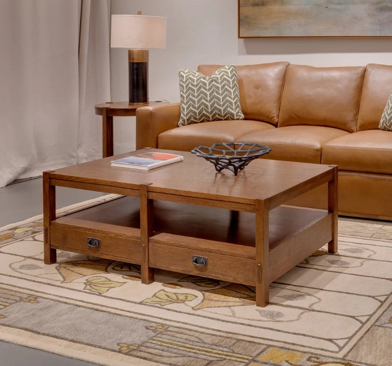 A Mission Style wooden coffee table with two drawers and an open shelf, styled in a living room next to a brown leather sofa on a patterned rug.
