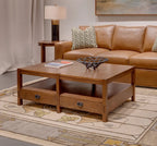 A Mission Style wooden coffee table with two drawers and an open shelf, styled in a living room next to a brown leather sofa on a patterned rug.