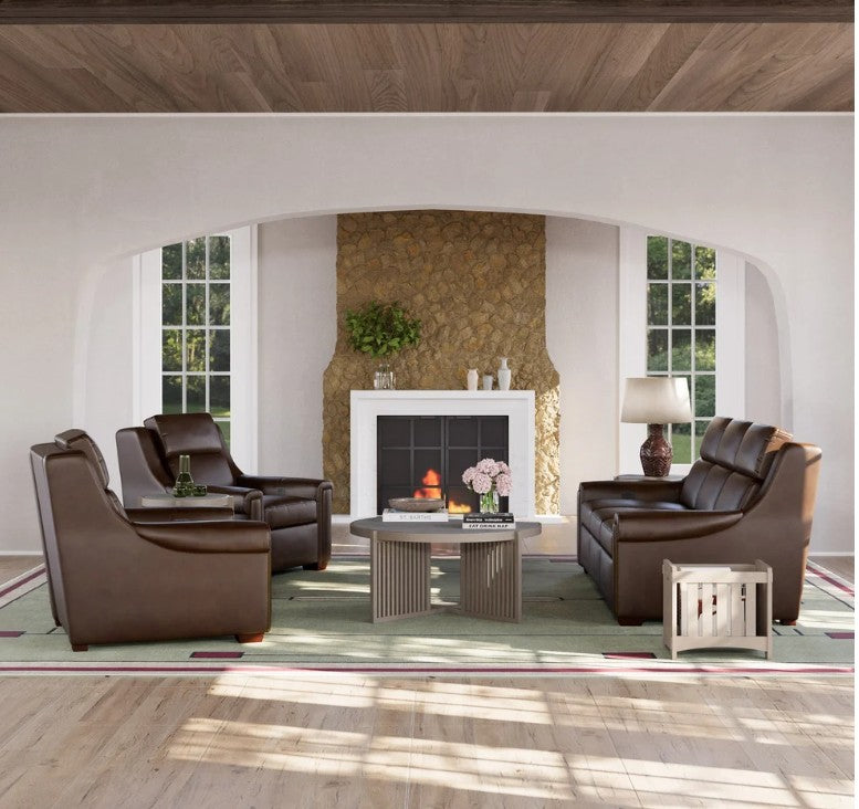 A styled living room featuring a dark brown leather club chair, matching armchair, and three-seater sofa arranged around a low ribbed coffee table on a green patterned rug. The room has a stone fireplace, arched windows, and a wood ceiling.