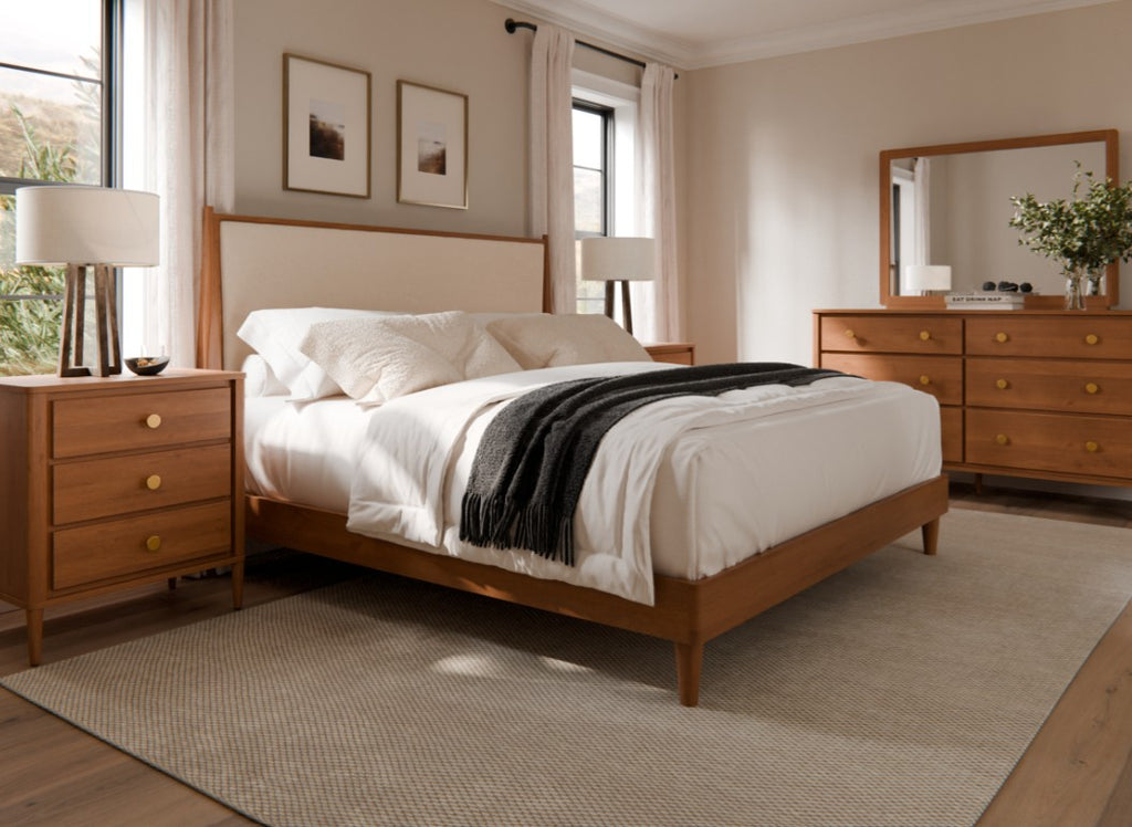 A warm, modern bedroom featuring a wooden bed frame with a cream upholstered headboard, styled with white bedding and a black throw blanket. The room includes matching wooden nightstands, a dresser, and a large mirror, all set against beige walls and a textured beige rug.