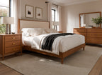 A warm, modern bedroom featuring a wooden bed frame with a cream upholstered headboard, styled with white bedding and a black throw blanket. The room includes matching wooden nightstands, a dresser, and a large mirror, all set against beige walls and a textured beige rug.