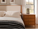 A modern bedroom scene featuring a wooden nightstand with three drawers and round brass knobs, styled next to a bed with white linens and a dark gray throw blanket. A white drum lamp with a dark wood tripod base sits on the nightstand, set against a window with sheer white curtains.