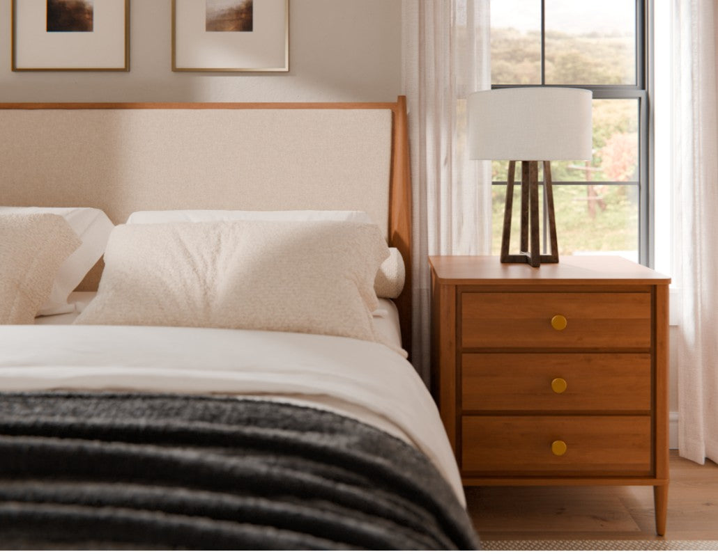 A modern bedroom scene featuring a wooden nightstand with three drawers and round brass knobs, styled next to a bed with white linens and a dark gray throw blanket. A white drum lamp with a dark wood tripod base sits on the nightstand, set against a window with sheer white curtains.