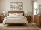 A styled bedroom featuring a wooden bed frame with cream bedding, matching wooden nightstands, a wooden dresser, and a large abstract painting on the beige wall.