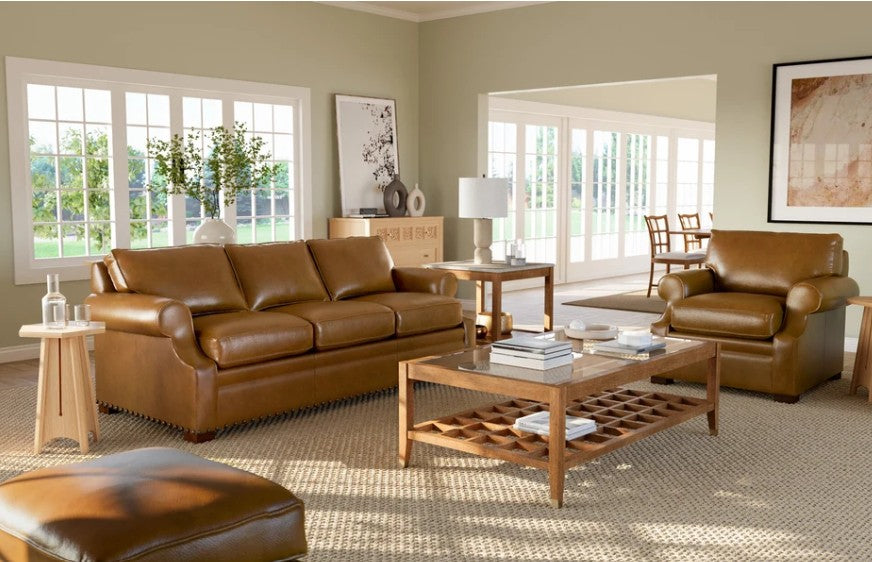 A brown leather three-seater sofa in a bright, modern living room with a matching armchair, wooden coffee table, and beige rug.