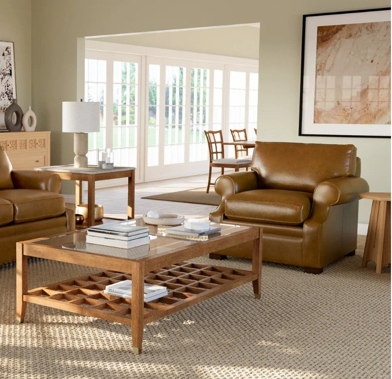 A brown leather armchair in a styled living room setting next to a wooden coffee table and a matching leather sofa, with a large window in the background.