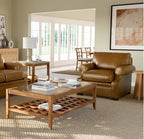 A brown leather armchair in a styled living room setting next to a wooden coffee table and a matching leather sofa, with a large window in the background.
