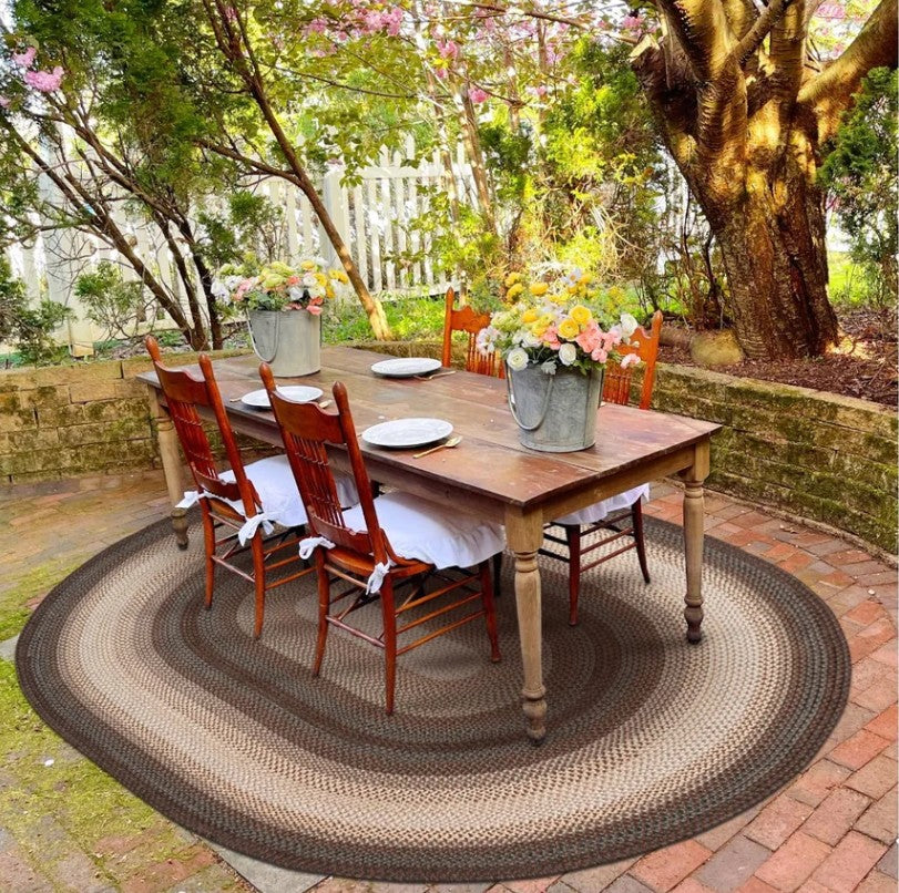 An outdoor dining scene with a rectangular wooden table set with six wooden chairs on a large round braided area rug. The table is decorated with white tablecloths, plates, and galvanized buckets of flowers.