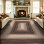 A large braided area rug with concentric rectangular patterns in shades of brown, beige, and gray, shown in a cozy living room setting with a fireplace, sofas, and windows.