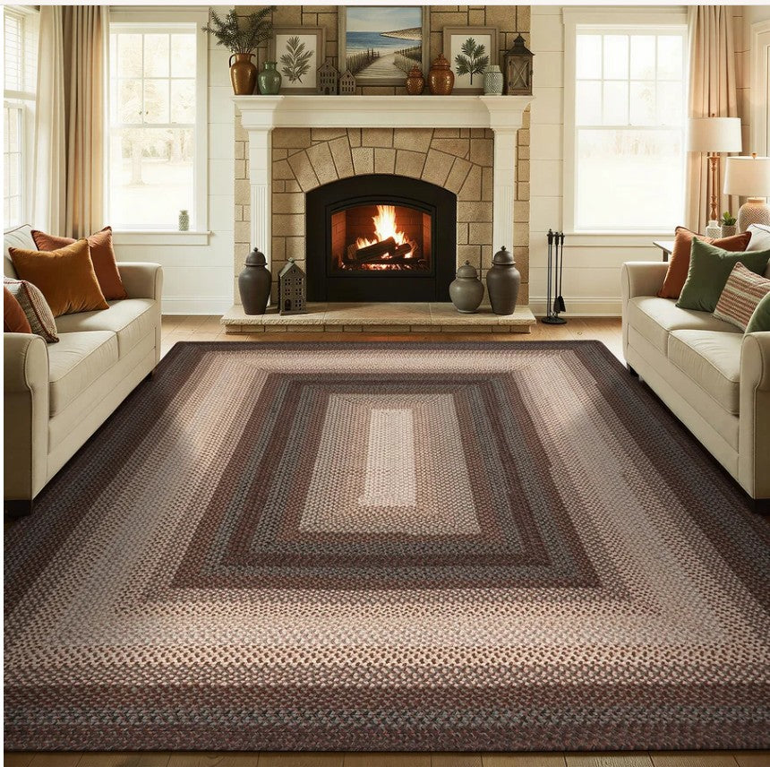 A large braided area rug with concentric rectangular patterns in shades of brown, beige, and gray, shown in a cozy living room setting with a fireplace, sofas, and windows.
