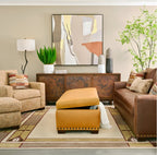 A styled living room featuring a mustard yellow ottoman with a lift-up top in the center, flanked by a beige armchair and a brown leather sofa. The room is decorated with a rusted wood sideboard, a large abstract painting, and a patterned area rug.