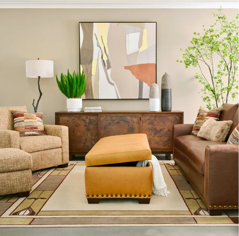 A styled living room featuring a mustard yellow ottoman with a lift-up top in the center, flanked by a beige armchair and a brown leather sofa. The room is decorated with a rusted wood sideboard, a large abstract painting, and a patterned area rug.