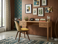 A mid-century modern wooden writing desk with three drawers and brass knobs, styled in a warm room with a yellow upholstered chair, framed botanical prints on a wood-paneled wall, and a brass floor lamp.