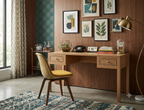 A mid-century modern wooden writing desk with three drawers and brass knobs, styled in a warm room with a yellow upholstered chair, framed botanical prints on a wood-paneled wall, and a brass floor lamp.