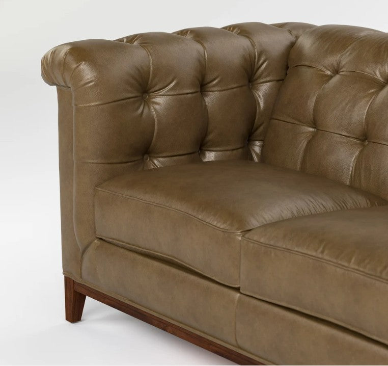 A close-up of a brown tufted leather sofa showing the backrest, armrest, seat cushion, and a portion of the wooden leg against a plain white background.