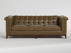 A classic Chesterfield-style leather sofa in a warm brownish-olive tone, featuring deep button tufting, rolled arms, and dark wooden legs, shown against a neutral light gray background.