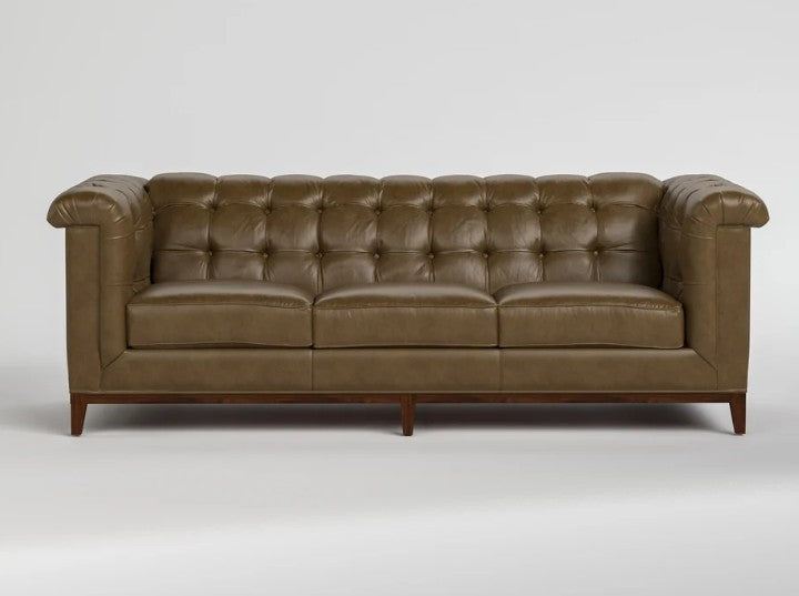 A classic Chesterfield-style leather sofa in a warm brownish-olive tone, featuring deep button tufting, rolled arms, and dark wooden legs, shown against a neutral light gray background.