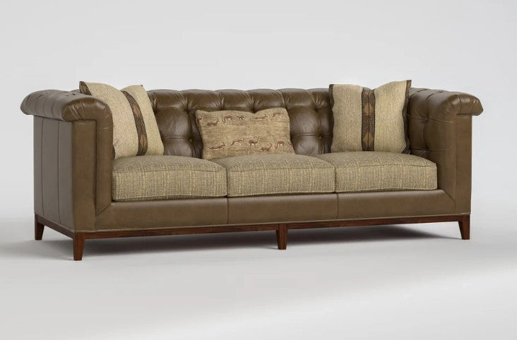 A classic Chesterfield-style sofa with brown leather tufted arms and back, upholstered in a textured beige fabric on the seat and back cushions, and accented with four matching beige throw pillows, shown on a light gray studio background.