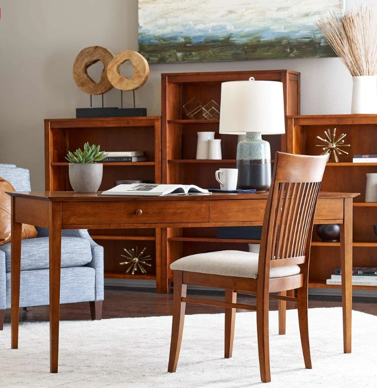 A mid-century modern wooden writing desk with a slatted back chair, styled in a warm, neutral-toned home office setting with a bookshelf, lamp, and decorative accents.