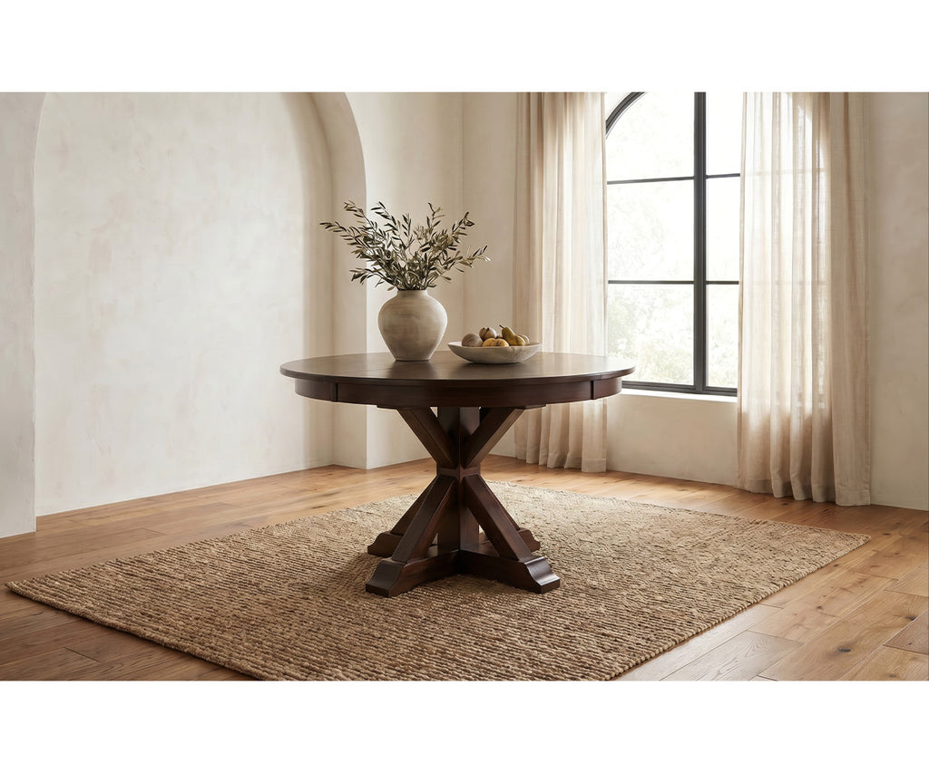 A round dark wood dining table with a cross base, styled on a woven jute rug in a bright room with beige walls, a window with sheer curtains, and a vase of branches on top.
