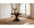A round dark wood dining table with a cross base, styled on a woven jute rug in a bright room with beige walls, a window with sheer curtains, and a vase of branches on top.