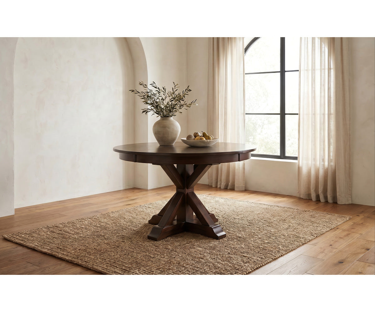 A round dark wood dining table with a cross base, styled on a woven jute rug in a bright room with beige walls, a window with sheer curtains, and a vase of branches on top.
