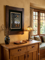A framed art print featuring a stylized tree and moon scene in earthy tones, displayed on a beige wall above a wooden console table in a warmly lit room.