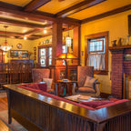 A warm, traditional interior room with wood ceiling beams, yellow walls, a brick fireplace, and a large wooden sofa with red cushions in the foreground.
