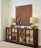Wooden sideboard with glass doors displaying decorative items, flanked by two lamps and a mirror.
