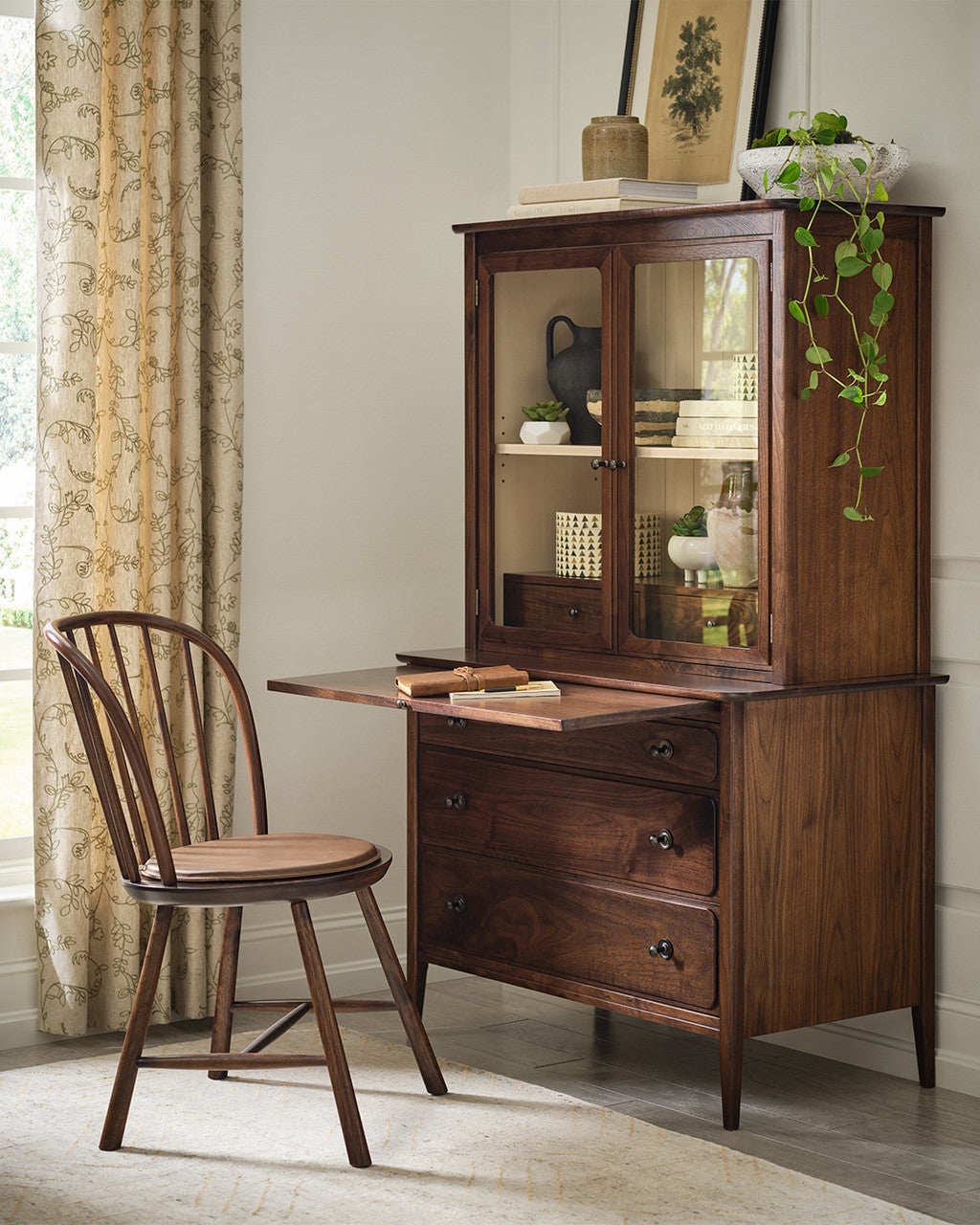 Wooden desk with glass cabinet and chair in a room with curtains and decor.