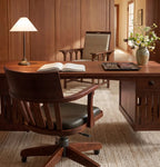 A warm wood office scene with a large wooden desk, a dark wood swivel chair with black leather seating, an open book on the desk, a table lamp, and a vase of flowers against wood-paneled walls.
