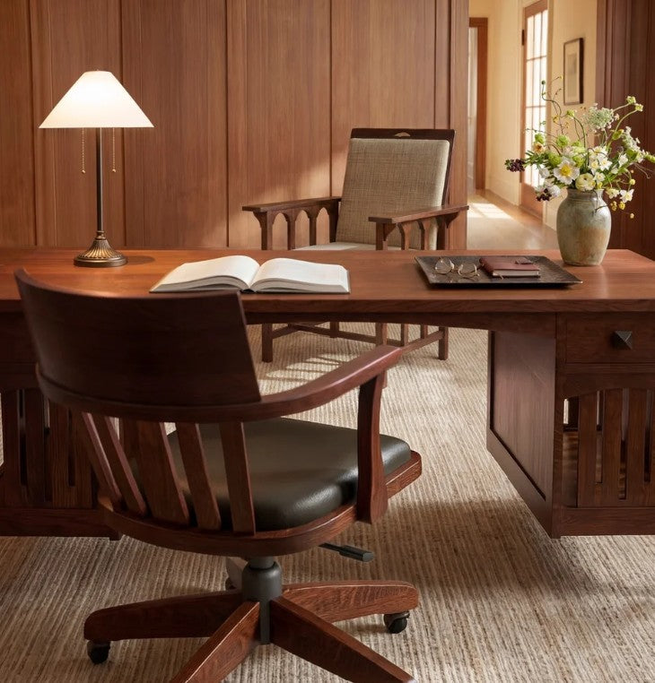 A warm wood office scene with a large wooden desk, a dark wood swivel chair with black leather seating, an open book on the desk, a table lamp, and a vase of flowers against wood-paneled walls.