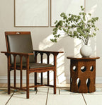 A mid-century modern wooden armchair with a dark brown wood frame and dark gray upholstered seat and back, positioned next to a matching wooden side table holding a white ceramic vase with green branches, set against a warm beige wall with framed art.
