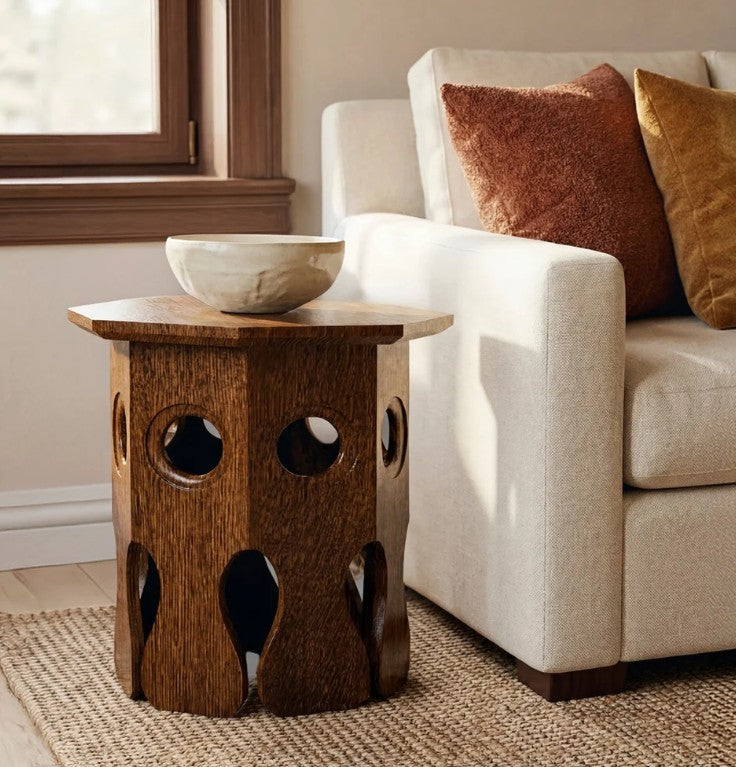 A rustic wooden side table with circular cutouts and a bowl on top, styled next to a cream-colored sofa with textured throw pillows in a warm, sunlit room.