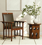 A mid-century modern wooden armchair with dark gray upholstered seat and backrest, positioned next to a matching wooden side table with a white ceramic vase of green branches, set against a neutral beige wall with framed art.